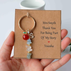 May include: A gold keyring with a red apple charm, gemstone beads, and a sparkling letter "S". The keyring is attached to a card that reads "Mrs Smyth Thank You For Being Part Of My Story".