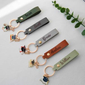 May include: Five personalized graduation keychains with names like "Sean" and "Brooke" on leather straps. Each keychain has a gold ring and a charm with a graduation cap, diploma, and the year 2026. The straps are in various colors.