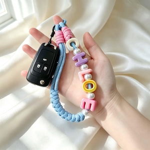 以下が含まれることがあります： 水色の編み込みコードキーホルダー。黒い車のキーフォブと、CHLOEという名前を綴るカラフルなプラスチック製の文字ビーズが特徴で、白い球状のビーズと上部のピンクのコードノットがアクセントになっています。