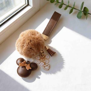 Puede incluir: Un juego de llaveros de cuero marrón con un pompón esponjoso, un charm con forma de Minnie Mouse con un lazo y una borla. El juego incluye una correa para la muñeca y un charm con las letras "CHAN" y un corazón con perlas.
