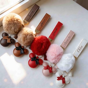 May include: A collection of personalized keychains featuring fluffy pom-poms and leather accents. Each keychain includes a leather tag with a name, and a Mickey Mouse-shaped leather charm. Colors include brown, red, pink, and white.