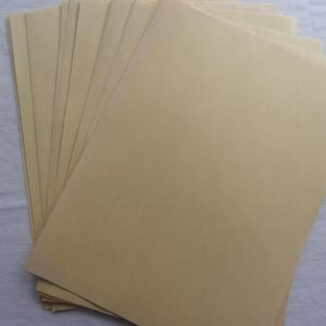 May include: A stack of tan-colored paper sheets, fanned out to display the edges. The paper has a smooth texture and a light brown hue. The sheets are arranged on a white surface.