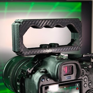 Può includere: Una fotocamera nera con una maniglia in fibra di carbonio attaccata. La fotocamera ha un obiettivo, un mirino e uno schermo che mostra un motivo a griglia verde. L'impugnatura ha una superficie strutturata e un'impugnatura curva.