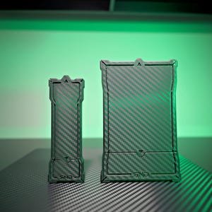 Può includere: Due custodie rettangolari nere con un motivo in fibra di carbonio. Le custodie hanno una superficie strutturata e un design unico. Le custodie sono su una superficie scura con uno sfondo verde.