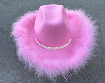 May include: A pink cowboy hat with a soft felt texture. The hat features a sparkling rhinestone band and is trimmed with fluffy pink feathers around the brim. A stylish accessory.