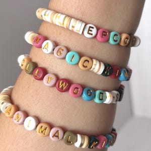 May include: Four colourful beaded bracelets spelling out "EPCOT", "MAGIC", "HOLLYWOOD", and "ANIMAL KINGDOM". The bracelets feature a variety of colours including pink, blue, white, and gold, with gold lettering on the beads.