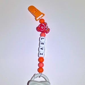 May include: A baby's dummy clip with the name "LEVI" spelled out in white letter beads. The clip features a red and orange cartoon fish charm, orange beads, and an orange clip. The dummy is grey.