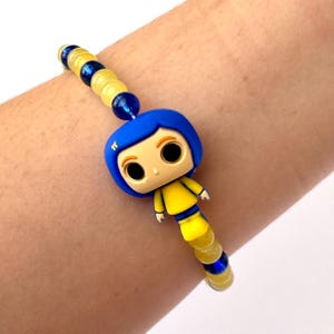 May include: A beaded bracelet featuring a blue and yellow figurine of a cartoon character. The bracelet has yellow and blue beads. The figurine has a blue bob hairstyle and a yellow outfit. The bracelet is worn on a wrist.
