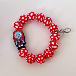May include: A red and white polka dot bead bracelet with a Sally charm. The bracelet features large, round beads with white polka dots. A Sally charm is attached, featuring a cartoon character with blue skin, red hair, and a patchwork dress. A silver-colored clasp is attached.