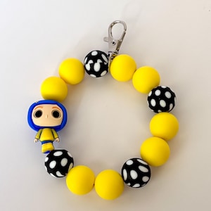 May include: A yellow and black beaded wristlet with a silver clasp. The wristlet features a small figurine of a character with blue hair and a yellow outfit. The beads are yellow and black with white polka dots.