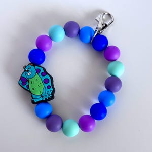 Puede incluir: Una pulsera de cuentas de colores con un dije de personaje de dibujos animados. La pulsera está hecha de cuentas azules, moradas y turquesas. Se adjunta un cierre plateado. El dije es un personaje de dibujos animados con pelaje azul, detalles morados y lunares verdes.