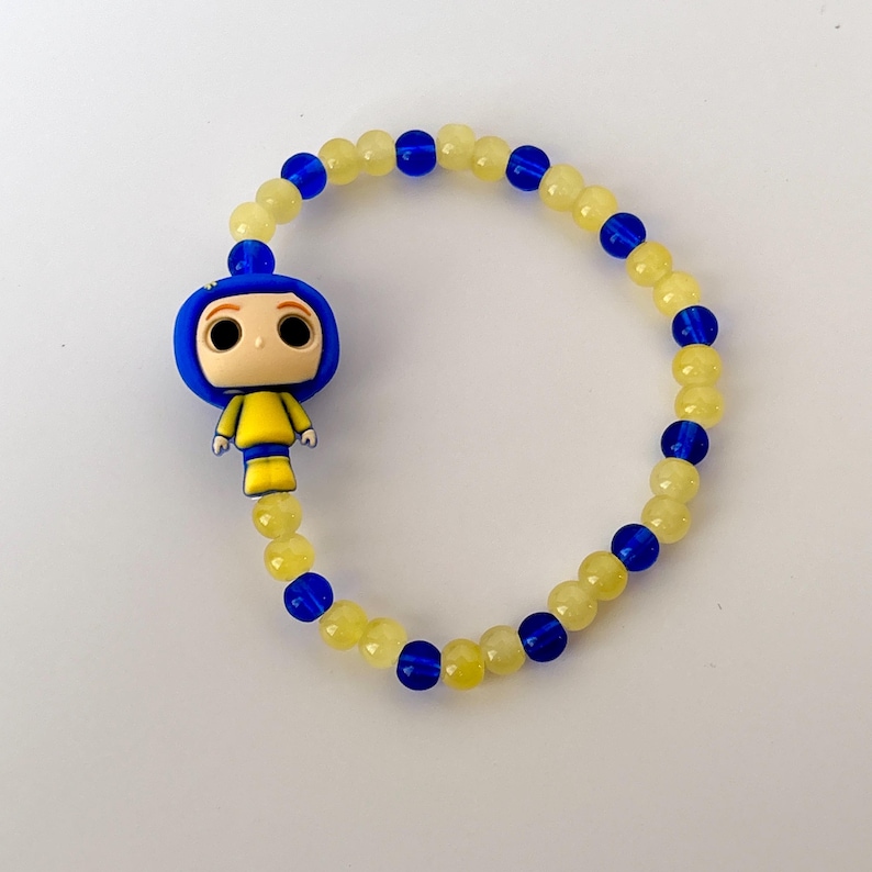 May include: A beaded bracelet featuring a cartoon character charm. The bracelet has yellow and blue beads, with a blue and yellow character charm. The bracelet is on a white surface.