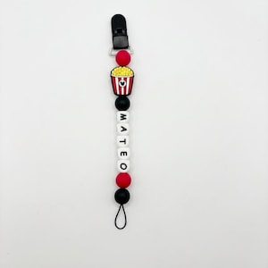 Puede incluir: Un sujetachupetes con una pinza de plástico negra, una cuenta roja, un dije de cubo de palomitas con rayas rojas y blancas, y cuentas de letras blancas que forman el nombre MATEO, junto con cuentas adicionales negras y rojas.
