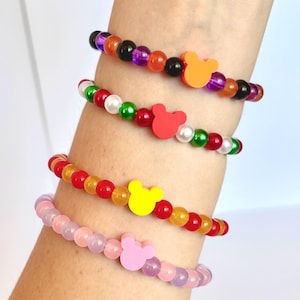 Puede incluir: Cuatro pulseras de cuentas con cuentas coloridas y dijes en forma de Mickey Mouse. Las pulseras presentan cuentas naranjas, negras, moradas, rojas, amarillas, verdes, blancas y rosas. Los dijes son naranjas, rojos, amarillos y rosas.