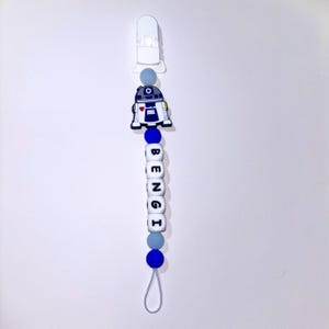 May include: A pacifier clip featuring a white plastic clasp at the top, followed by a blue, white, and grey color scheme. It includes a silicone robot character charm and white cube beads spelling the name BENGI in black letters.