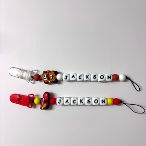 May include: Two baby pacifier clips with the name "JACKSON" spelled out in white letter beads. One clip has a red and yellow car charm, the other has a brown car charm. Both have a clear pacifier.