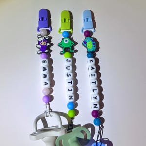 Peut inclure: Trois attaches-sucettes personnalisées avec des breloques de personnages. Chaque attache a une pince et un charme de couleur différente, avec des noms épelés en perles de lettres blanches : Emma, Justin et Kaitlyn. Les sucettes sont vert clair et grises.