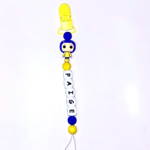 May include: A pacifier clip featuring a yellow plastic clasp at the top, followed by a blue and yellow character figurine, and a series of white spherical beads with black letters spelling out the name PAIGE, finished with blue and yellow beads.