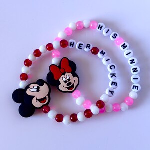 May include: Two beaded bracelets featuring Mickey and Minnie Mouse charms. One bracelet spells "HER MICKEY" and the other spells "HIS MINNIE". The beads are white, pink, and red.