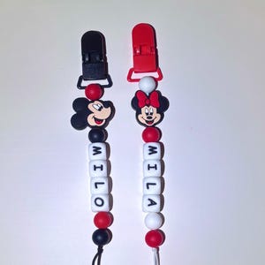 May include: Two pacifier clips featuring Mickey and Minnie Mouse. One clip has a black clip and Mickey Mouse, the other has a red clip and Minnie Mouse. Each clip has the name "MILA" spelled out in white letter beads.