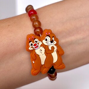 May include: A beaded bracelet featuring a charm of Chip and Dale. The charm is orange and tan with black accents. The beads are brown, red, and amber. The bracelet is worn on a wrist.