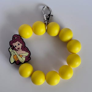 May include: Yellow beaded keychain with a silver clasp and a cartoon image of a woman with long brown hair, wearing a yellow dress. The keychain is made of yellow beads and is a fun accessory.