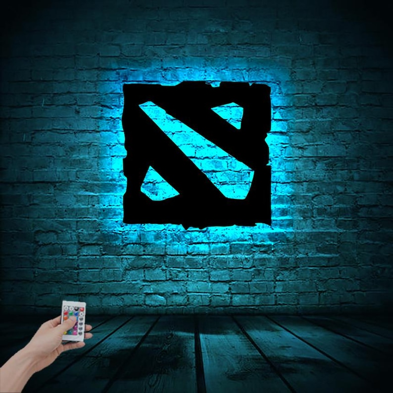 Puede incluir: Una luz azul ilumina un logotipo de Dota 2 en blanco y negro en una pared de ladrillo. Un mando a distancia con botones de colores est&aacute; en primer plano.