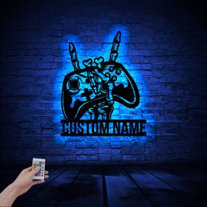 May include: A blue neon sign with a video game controller design. The controller is holding up two fingers in a rock and roll hand gesture. The sign has a space for a custom name below the controller.