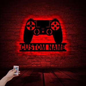 May include: A black silhouette of a video game controller with the text "CUSTOM NAME" underneath. The controller is illuminated in red and mounted on a brick wall. A remote control is visible in the bottom left corner of the image.