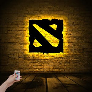 Puede incluir: Un letrero iluminado negro y amarillo con el logotipo de Dota 2 en una pared de ladrillo. El letrero est&aacute; iluminado con un brillo amarillo y se controla con un mando a distancia.