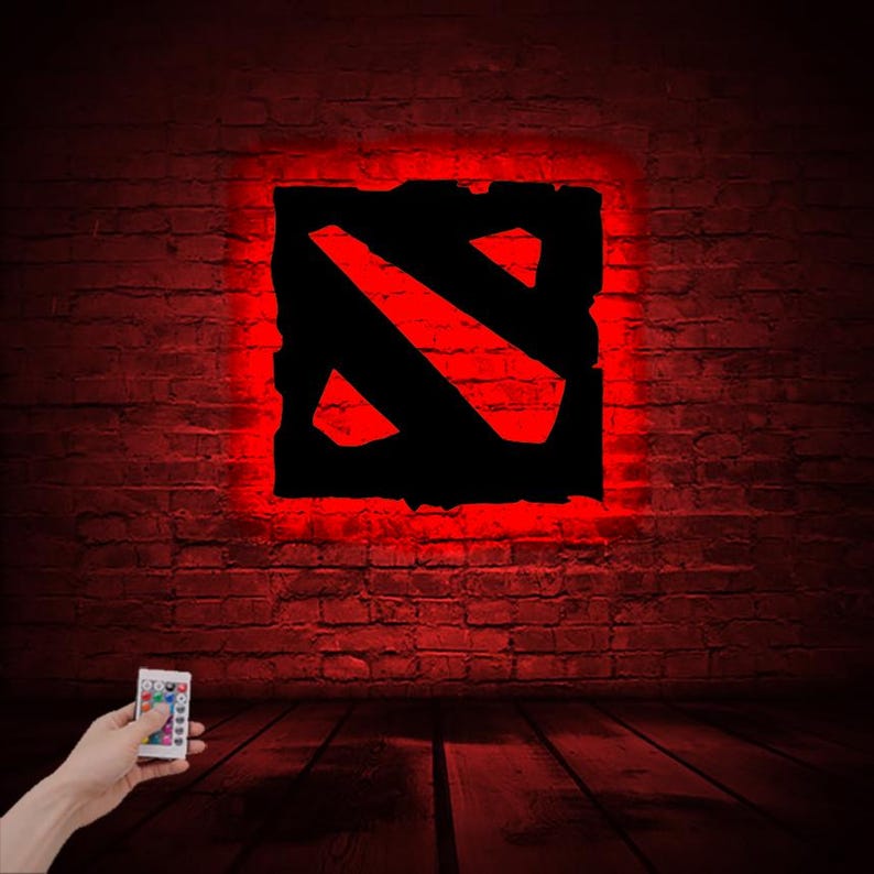 Puede incluir: Una luz roja y negra con el logotipo de Dota 2 y un mando a distancia. La luz est&aacute; sobre un fondo de pared de ladrillo.