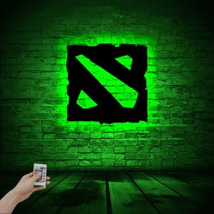 Puede incluir: Un letrero luminoso verde y negro con el logotipo de Dota 2. El letrero est&aacute; en una pared de ladrillo y est&aacute; iluminado con luz verde. Un mando a distancia est&aacute; en primer plano.