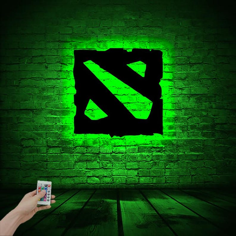 Puede incluir: Un letrero luminoso verde y negro con el logotipo de Dota 2. El letrero est&aacute; en una pared de ladrillo y est&aacute; iluminado con luz verde. Un mando a distancia est&aacute; en primer plano.
