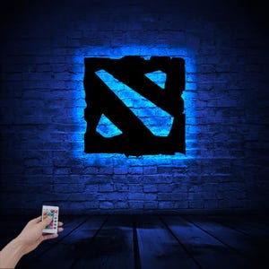 Puede incluir: Un letrero de ne&oacute;n negro y azul con el logotipo de Dota 2 en una pared de ladrillo. El letrero est&aacute; iluminado con luz azul y tiene un control remoto.