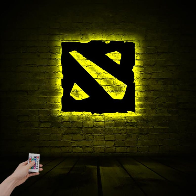 Puede incluir: Un letrero de ne&oacute;n amarillo con el logotipo de Dota 2 en una pared de ladrillo. El letrero est&aacute; iluminado y viene con un control remoto.