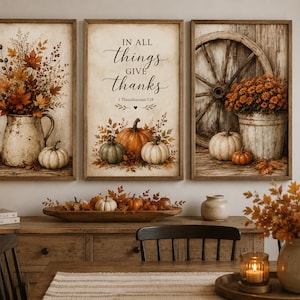 May include: A set of three rectangular wall art prints featuring autumn themes. The left print shows a pitcher with fall foliage and a pumpkin. The center print displays the text IN ALL THINGS GIVE THANKS with pumpkins. The right print shows a wagon wheel.