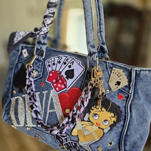May include: A denim handbag with various patches, including playing cards, dice, and a Betty Boop design. The bag features a chain strap, a black and white patterned scarf tied to the handle, and the name "Oliva" in rhinestones.