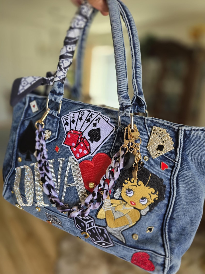 May include: A denim handbag with various patches, including playing cards, dice, and a Betty Boop design. The bag features a chain strap, a black and white patterned scarf tied to the handle, and the name "Oliva" in rhinestones.