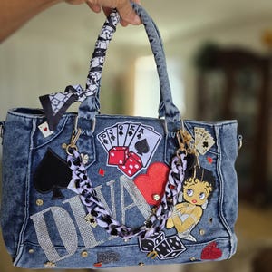 May include: A blue denim handbag with a variety of patches and embellishments. The bag features playing card suits, dice, and a Betty Boop patch. The word "DIVA" is spelled out in rhinestones. The handles are wrapped in a black and white patterned fabric.
