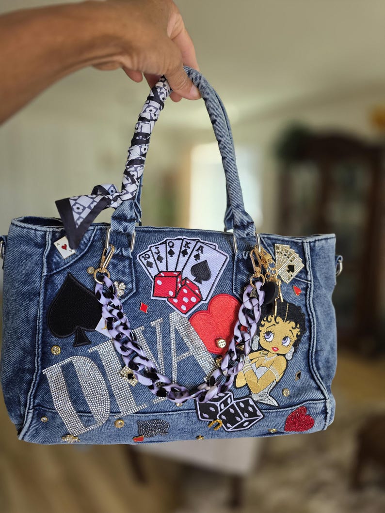 May include: A blue denim handbag with a variety of patches and embellishments. The bag features playing card suits, dice, and a Betty Boop patch. The word "DIVA" is spelled out in rhinestones. The handles are wrapped in a black and white patterned fabric.
