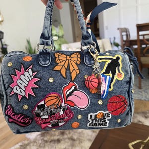 Puede incluir: Bolso de mano de mezclilla con varios parches, incluyendo un bocadillo rosa "BAM!", un lazo naranja y dise&ntilde;os tem&aacute;ticos de baloncesto. El bolso cuenta con un asa superior, un llavero con dijes de baloncesto y tachuelas decorativas.