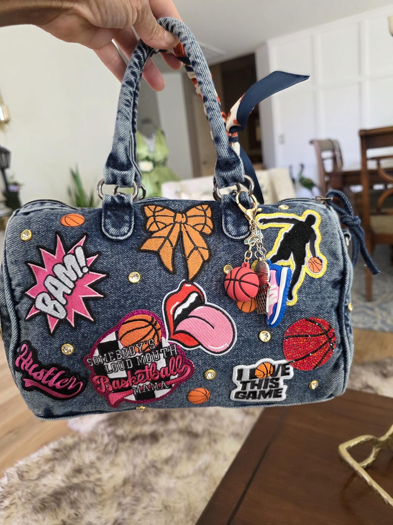 Puede incluir: Bolso de mano de mezclilla con varios parches, incluyendo un bocadillo rosa "BAM!", un lazo naranja y dise&ntilde;os tem&aacute;ticos de baloncesto. El bolso cuenta con un asa superior, un llavero con dijes de baloncesto y tachuelas decorativas.