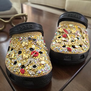 Puede incluir: Un par de sandalias Crocs negras, muy adornadas con pedrería dorada, plateada e iridiscente. Las sandalias están adornadas con abalorios que incluyen labios, notas musicales y corazones. Las suelas son negras.