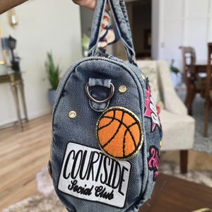 Puede incluir: Un bolso de mezclilla con parches y adornos. El bolso presenta un parche de baloncesto, el texto "COURTSIDE Social Club" y otros elementos decorativos. El bolso tiene un asa y una correa con una bufanda estampada.