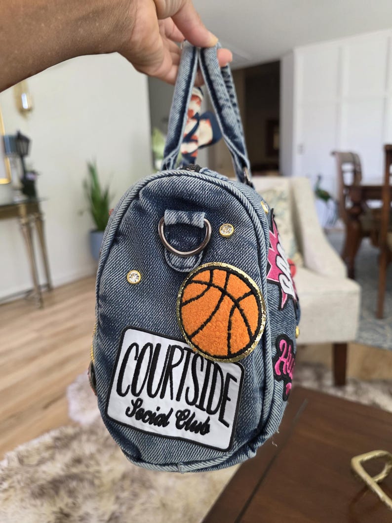 Puede incluir: Un bolso de mezclilla con parches y adornos. El bolso presenta un parche de baloncesto, el texto "COURTSIDE Social Club" y otros elementos decorativos. El bolso tiene un asa y una correa con una bufanda estampada.