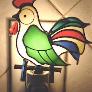 May include: A stained glass night light shaped like a rooster. The rooster features a red comb and wattle, a green body, and a multi-colored tail. The night light plugs directly into a wall outlet.