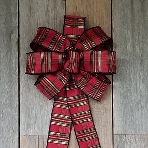 May include: A decorative red plaid bow with gold accents and black trim. The bow is made of a wide ribbon and has long, flowing tails. The bow is likely for holiday or gift decoration.