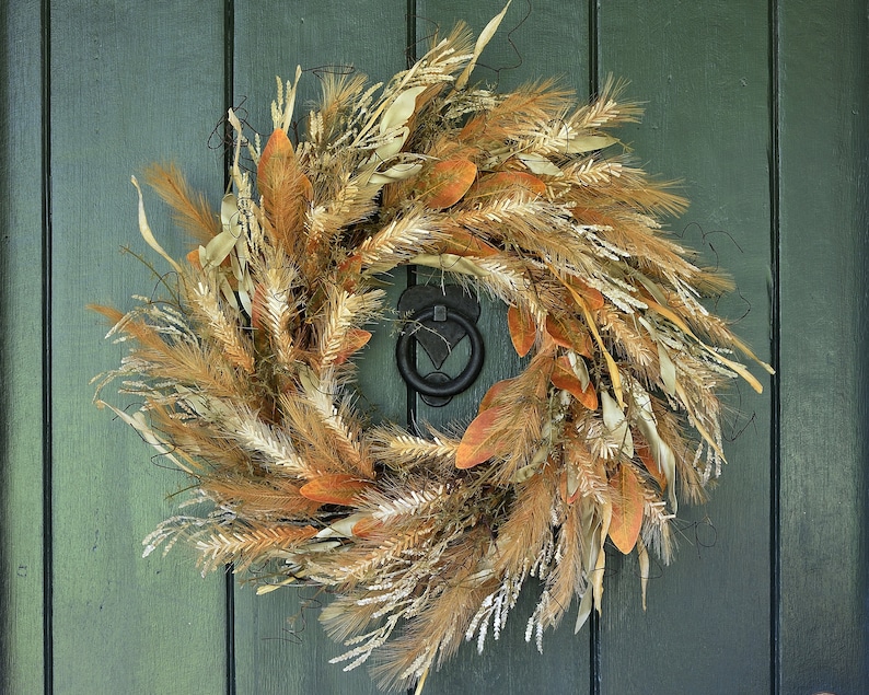 May include: A wreath made of dried grasses, wheat, and leaves in shades of brown, beige, and white. The wreath is hanging on a dark green door.