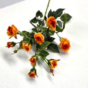 May include: A spray of artificial flowers featuring multiple small, vibrant orange rose blossoms and buds. The stems are thin and green, accompanied by several dark green, serrated leaves arranged along the branches of the floral arrangement.