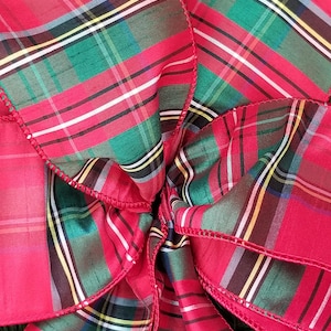 May include: Close-up of a large, red plaid bow with green, black, white, and yellow accents. The bow is made of a shiny fabric and has a decorative, stitched edge. The bow is likely for holiday or gift decoration.
